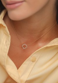 Collar de cadena dorada con un colgante en forma de flor, incrustado con piedras transparentes. Llevado sobre una camisa de botones de color amarillo pálido.