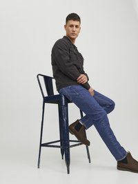 Černá džínová bunda s předními kapsami, modré dž�íny a hnědé kotníkové boty. Osoba sedí na opotřebované modré kovové barové židli.