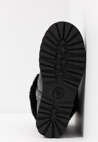 Semelle noire de botte avec un motif de bande de roulement profond et un logo circulaire, reposant contre un mur blanc.