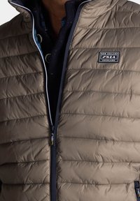 Beige puffer gilet met gestikte textuur, frontale ritssluiting, marineblauwe accenten en geborduurd logo. Heeft een hoge kraag en een binnenlaag van stof.