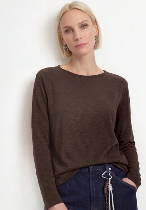 Mujer rubia con una camiseta marrón de manga larga y vaqueros azul oscuro con un cinturón de cuerda, de pie con una mano en el bolsillo sobre un fondo blanco.