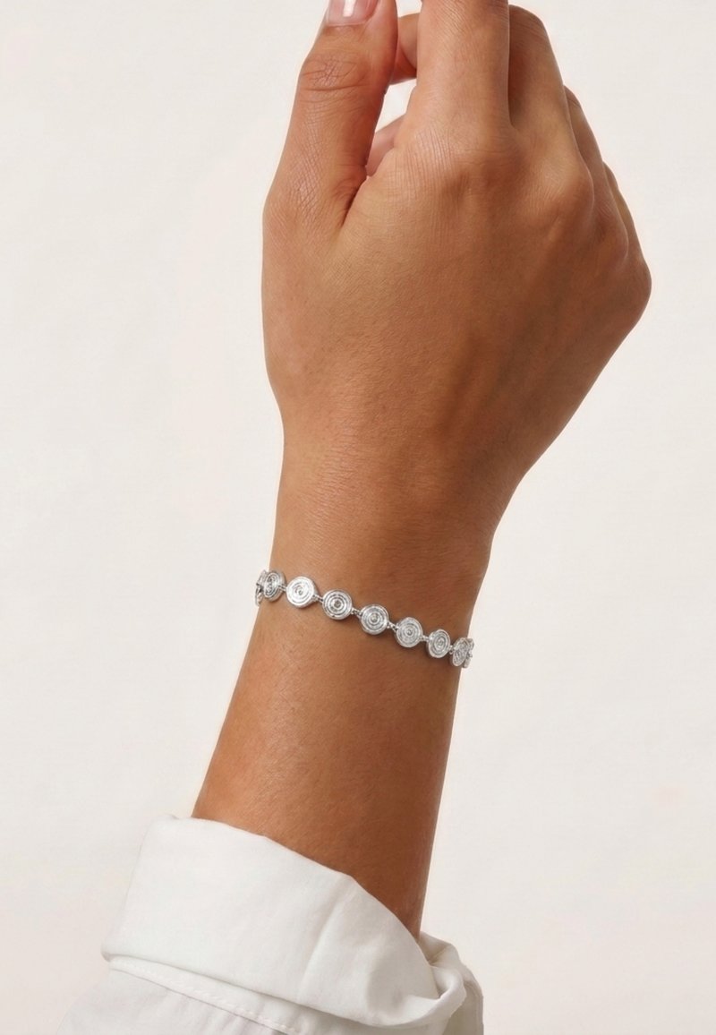 Bras avec un bracelet en argent composé de maillons circulaires en spirale, sur un fond clair uni et un poignet de chemise blanc.