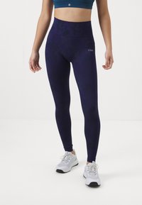 Marineblauwe high-waisted leggings van een soepele, rekbare stof met een subtiele patroon; gecombineerd met witte sportschoenen en een teal sporttop.