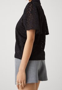 Svart spetsblus med korta ärmar, med ett blommönster och en solid krage. Matchad med grå shorts som har en avslappnad passform.