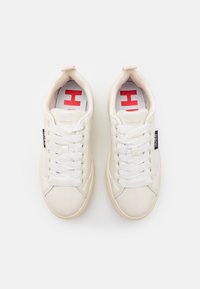 Zapatillas blancas de cuero con textura suave, puntera redonda, cordones planos y detalle del logotipo en contraste en el lateral. Interior con un patrón rojo visible.