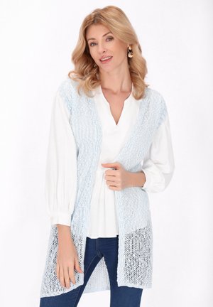 Blond vrouw draagt een witte blouse, lichtblauw gebreid mouwloos vest en blauwe spijkerbroek, glimlachend tegen een witte achtergrond.