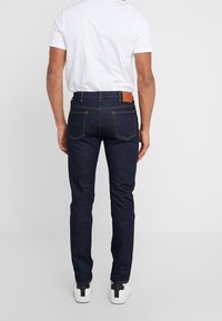 Muž oblečený v tmavě modrých úzkých džínách, bílém tričku a bílých teniskách, stojící zády k nám na jednoduchém bílém pozadí.