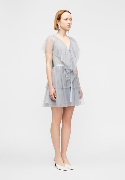 Νεαρή γυναίκα που φοράει ένα ανοιχτό γαλάζιο φόρεμα από τούλι με στρώσεις και βολάν, μακριά σκουλαρίκια με πέρλες και λευκά παπούτσια μυτερά με χαμηλό τακούνι, στέκεται μπροστά σε λευκό φόντο.