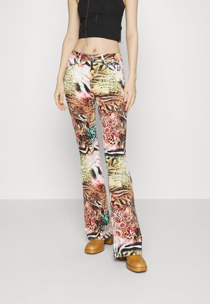 Nainen pukeutuneena värikkäisiin eläinkuosisiin liehuhousuihin, mustaan hihattomaan lyhythihaiseen paitaan ja beigeihin korotettuihin kenkiin, seisomassa yksinkertaisen vaalean taustan edessä.