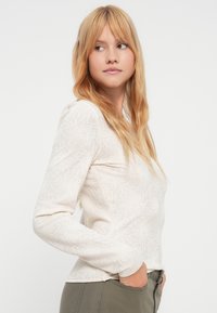 Κρεμ μακρυμάνικο τοπ με φλοράλ υφή, στρογγυλή λαιμόκοψη και εφαρμοστή σχεδίαση. Συνδυασμένο με παντελόνι λαδί, με τα χέρια να ακουμπούν στις τσέπες.