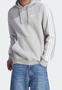 Grå hoodie med tonad design, som har en känguruficka och vita tre-stripes detaljer på ärmarna. Mjuk material med dragsko i huvan.