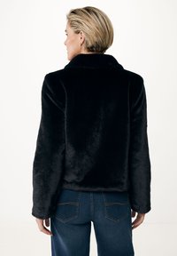Svart faux fur-jacka med krage, mjuk textur och avslappnad passform. Modellen bär blå jeans med en klassisk bakfickedesign.