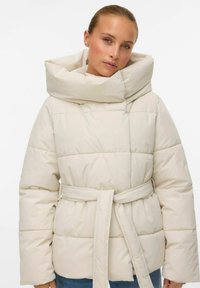 Heledbeez puffer-jakk, millel on lai, volditud krae ja vöökoht. Omab tikandit ja pikki varrukaid.