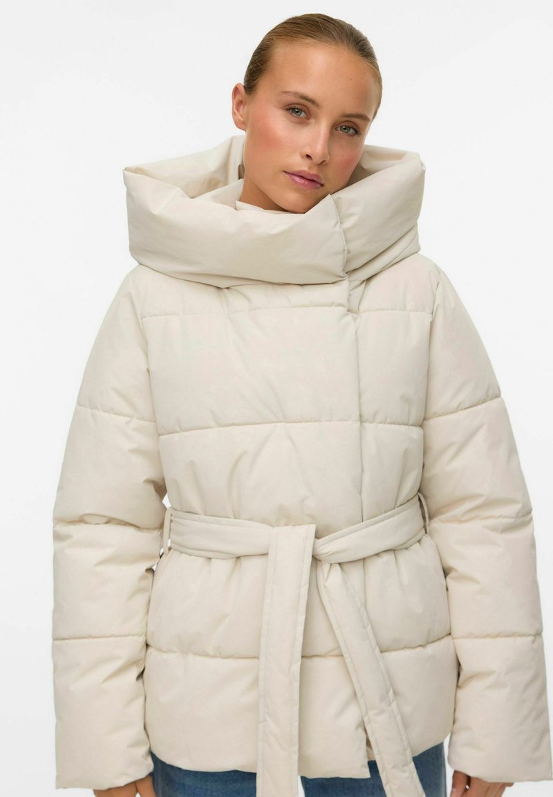 Heledbeez puffer-jakk, millel on lai, volditud krae ja vöökoht. Omab tikandit ja pikki varrukaid.