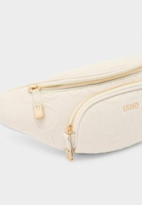 Creme faux læder bæltetaske med to guld lynlåskomponenter, præget logo mønster og en guld hardware detalje på lynlås trækkeren.