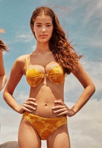 Gele bikini met bloemenpatroon, voorzien van een gepadde, gestructureerde top met een strik in het midden en een bijpassende low-rise onderkant, gemaakt van een gladde stof.