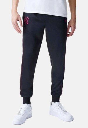 Mann trägt schwarze Jogginghose mit rotem NY-Logo und roten Seitenstreifen, weiße Sneaker und ein weißes Hemd, steht vor einem einfarbigen Hintergrund.