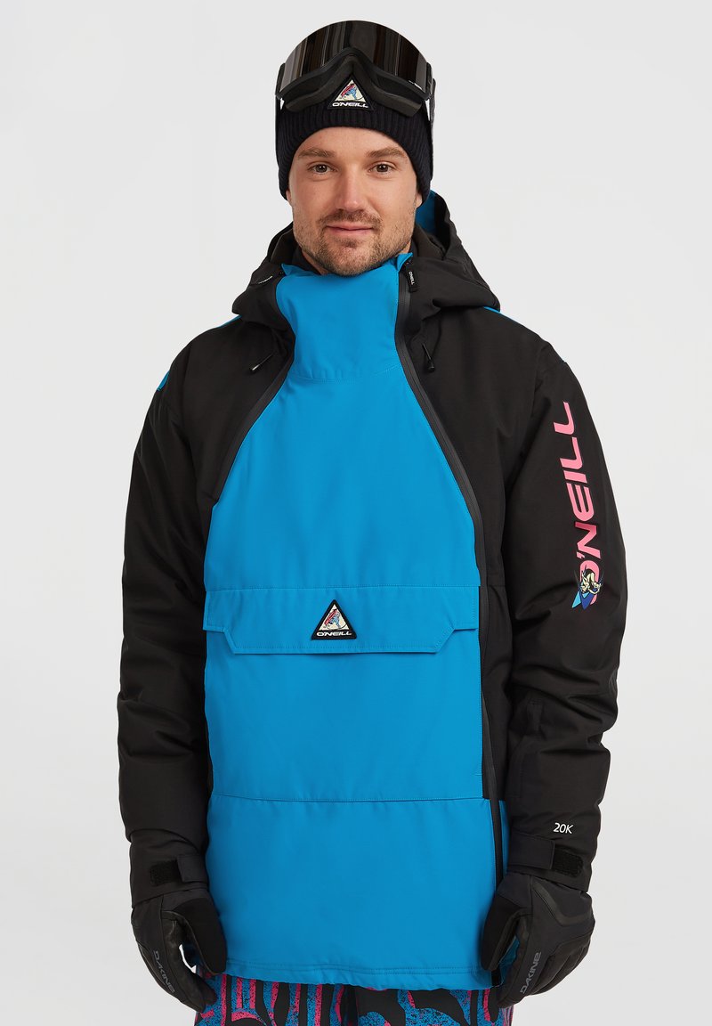 Chaqueta de nieve azul y negra con un logo triangular, bolsillo frontal y texto rosa en la manga. Se llevan guantes negros y gafas de esquí.