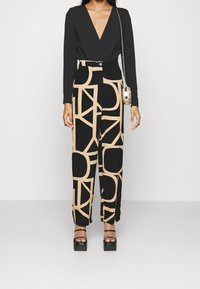 Kobieta w czarnej bluzce z głębokim dekoltem w serek, w czarnych spodni wysokiej talii z dużym beżowym geometrycznym wzorem, na platformach oraz z małą okrągłą torebką na łańcuszku.