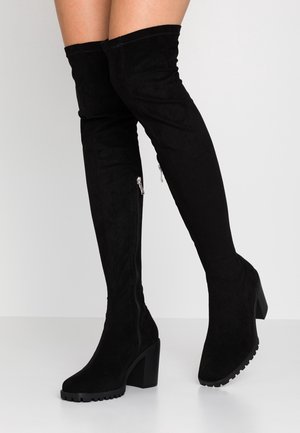 Bottes hautes en daim noir au-dessus du genou avec talons blocs et fermetures éclair latérales, portées par une personne debout devant un fond clair uni.