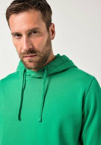 Vihreä huppari, joka on valmistettu pehmeästä kankaasta. Hupparissa on kiristysnauha hupussa, resorireunukset hihoissa ja sileä pinta ilman näkyviä kuvioita tai logoja.