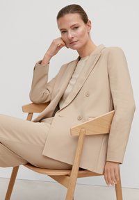 Beige Doppelreiher-Anzug mit taillierter Passform, glattem Stoff und zwei Knöpfen. Das Model sitzt auf einem Holzstuhl vor einem neutralen Hintergrund.