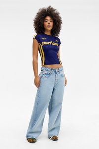 Tânără cu părul creț, purtând un top scurt bleumarin cu dungi galbene și blugi largi de culoare albastru deschis, stând pe un fundal alb simplu.