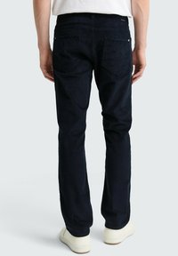 Granatowe sztruksowe spodnie wyświetlone od tyłu, z dwoma tylnymi kieszeniami i teksturowaną tkaniną. Noszone z jasnymi sneakersami.