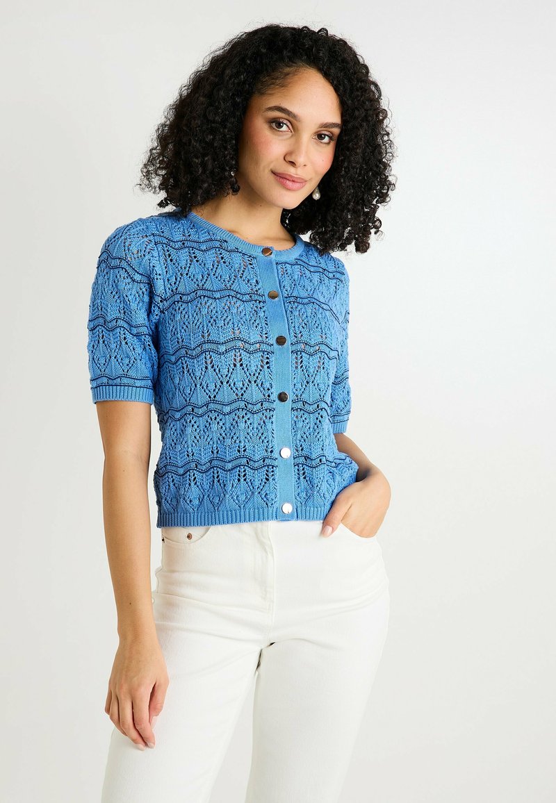 Cárdigan de punto azul con un diseño de encaje, mangas cortas y cierre frontal con botones. Presenta un dobladillo acanalado y patrones texturizados. Combinado con pantalones blancos.