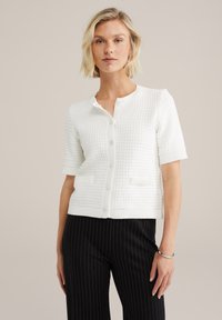Bílý cardigan s krátkým rukávem a texturovaným vzorem, s knoflíkovým zapínáním a dvěma předními kapsami. Má kulatý výstřih a žebrované manžety.