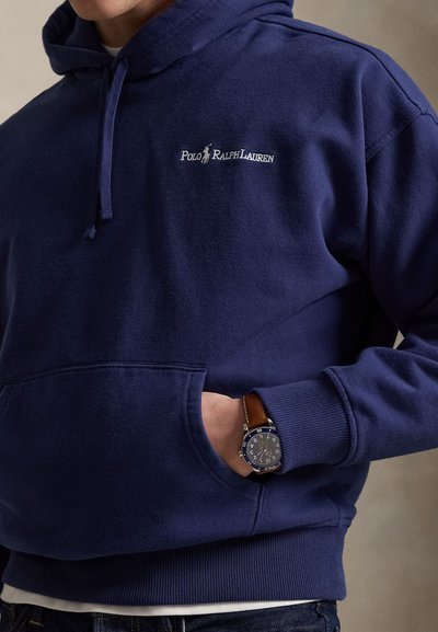 Muž v tmavomodrej mikine Polo Ralph Lauren s rukou vo vrecku na prednej strane a hnedými koženými hodinkami na zápästí.