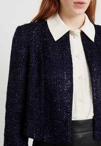 Námořnický tweedový sako s texturovaným povrchem, s bílým límcem, zkráceným střihem a jemnými metalickými nitěmi propletenými po celém povrchu.