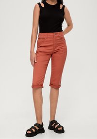 Pantaloni cropped color ruggine con risvolti, design a cinque tasche e top senza maniche nero. Calzature sono sandali neri chunky.