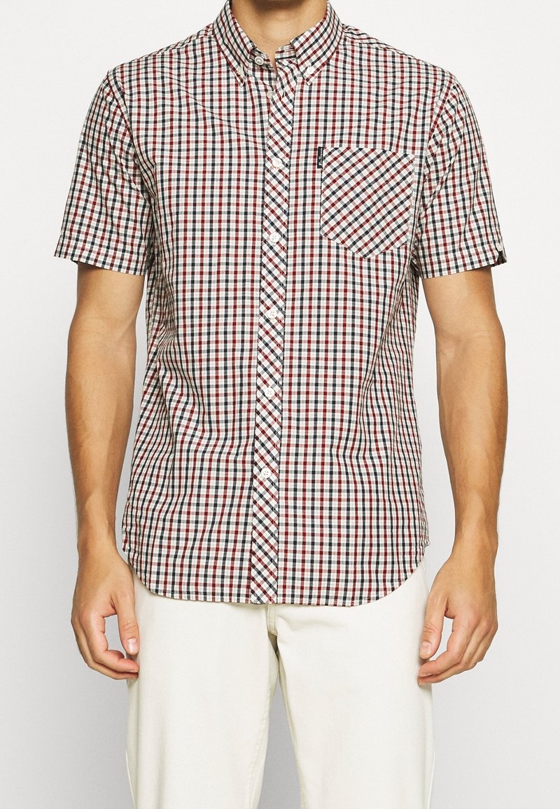 Camisa de botones de manga corta en un patrón a cuadros rojo, blanco y negro. Cuenta con un bolsillo en el pecho y un diseño de cuello clásico.