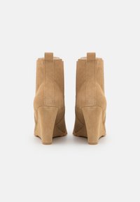 Botas de tornozelo em camurça bege com salto alto, bico redondo e alça traseira. A superfície tem uma textura suave e costuras visíveis.