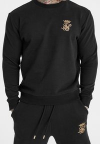 Sudadera negra con un logotipo bordado en dorado en el pecho, puños y dobladillo acanalados. Tejido suave con un corte relajado.