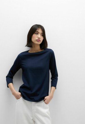 Frau mit glatten dunklen Haaren, die ein marineblaues Langarmshirt und weiße Hosen trägt, mit den Händen in den Taschen stehend vor einer schlichten weißen Wand.