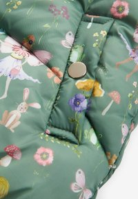 Tejido verde acolchado con patrones florales y de animales, presenta un cierre de botón a presión plateado y costuras en contraste, textura suave.