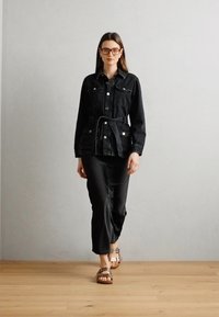 Jachetă din denim negru cu talie cu curea, închidere cu nasturi și două buzunare pe piept, purtată cu pantaloni negri și sandale maro. Podea din lemn texturat.