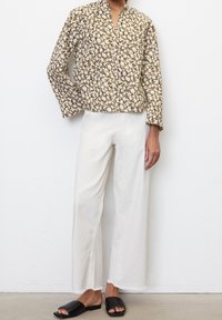 Blusa marrone a maniche lunghe con motivo floreale e colletto alla mandarina, abbinata a pantaloni bianchi a gamba larga e sandali neri.