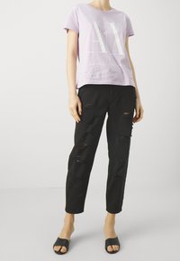 Camiseta de manga corta lila con el logo "AX", combinada con jeans negros desgastados con múltiples desgarros. Las sandalias negras de tipo slide completan el look.