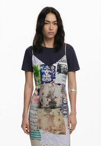Mehrfarbiges Patchwork-Kleid mit Spaghettiträgern, das über einem dunklen T-Shirt getragen wird. Es weist abwechslungsreiche Muster, Texturen und Drucke in Blau-, Grün- und Grautönen auf.
