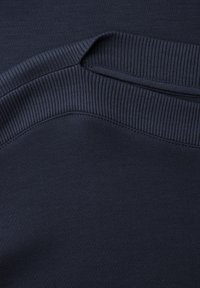 Detalje af mørk marineblåt ribstrikket stof, der viser kraven og skuldersømmen på et beklædningsgenstand, med fokus på struktur og syning.