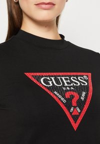 Schwarzes T-Shirt mit kurzen Ärmeln, das ein auffälliges rotes und schwarzes dreieckiges Logo mit weißem Text und einem zentralen Fragezeichen-Design zeigt.
