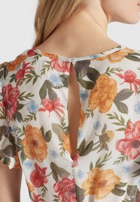 Rückansicht einer Person, die eine Bluse mit Blumenmuster in Orange, Rot und Blau trägt, mit einem Schlüsselloch-Ausschnitt hinten und einem kleinen Knopfverschluss.