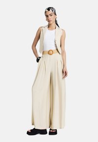 Un gilet leggero beige, corto, indossato sopra una canotta bianca, abbinato a pantaloni larghi a vita alta. Presenta una cintura intrecciata con una grande fibbia rotonda.