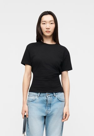 Mlada ženska s ravnimi temnimi lasmi, oblečena v oprijeto črno majico in svetlo modre kavbojke, drži ob strani majhno črno torbico.