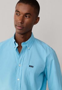 Camisa azul claro de algodón con botones, con un bolsillo en el pecho izquierdo y una pequeña etiqueta de la marca. Diseño de cuello clásico, textura suave.