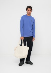 Blå crewneck sweatshirt med en liten gul logo, kombinert med mørke bukser og en stor kremfarget totebag med en teksturert frontlomme.