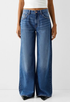 Kvinde iført højtaljede, viddebenede blå jeans med falmet detaljering, kombineret med sorte spidse sko og en kort hvid top.